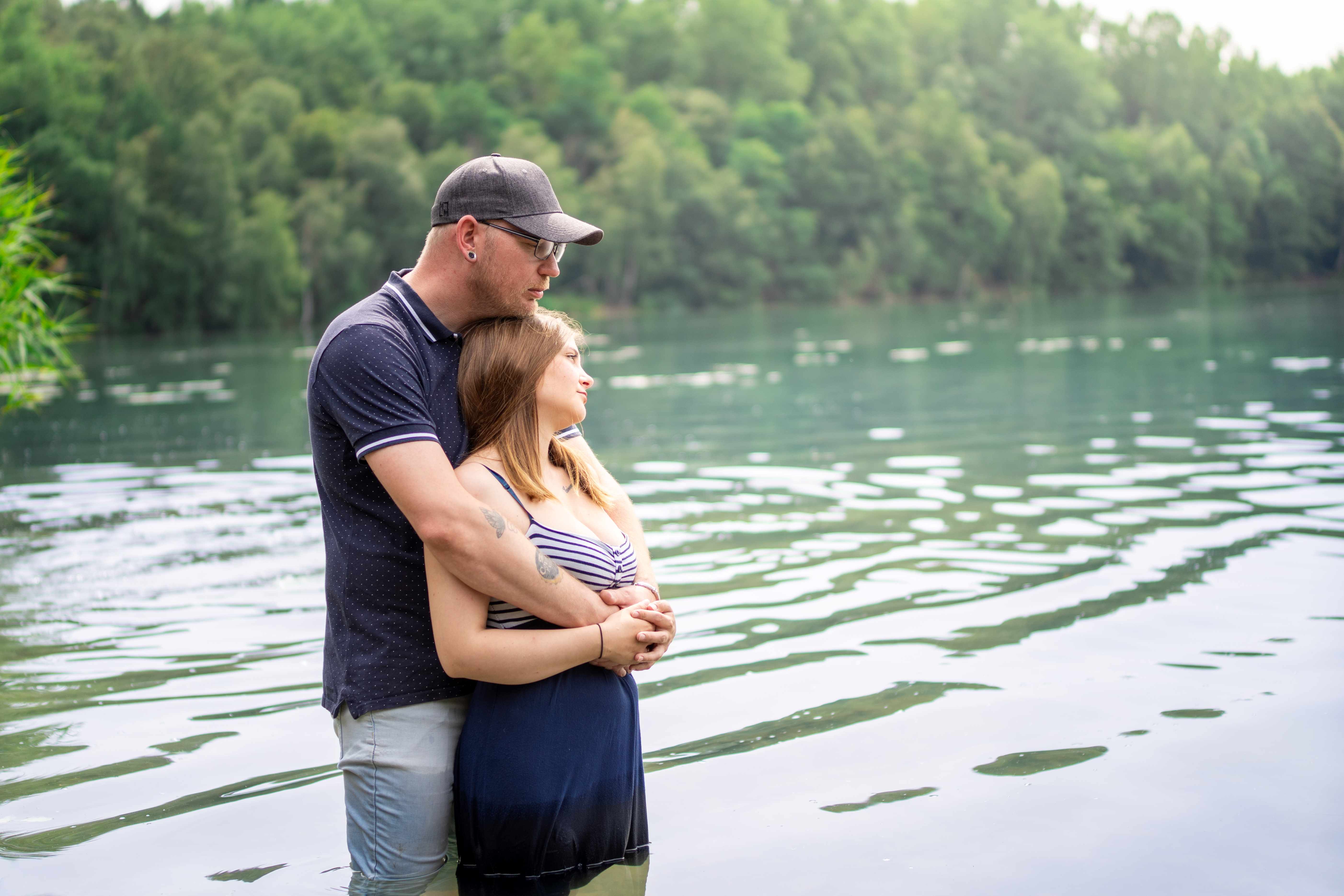 Paar steht im Wasser am See, der Mann hält die Frau liebevoll von hinten im Arm. Sie schauen gemeinsam in die Ferne, umgeben von ruhigem Wasser und grüner Natur.