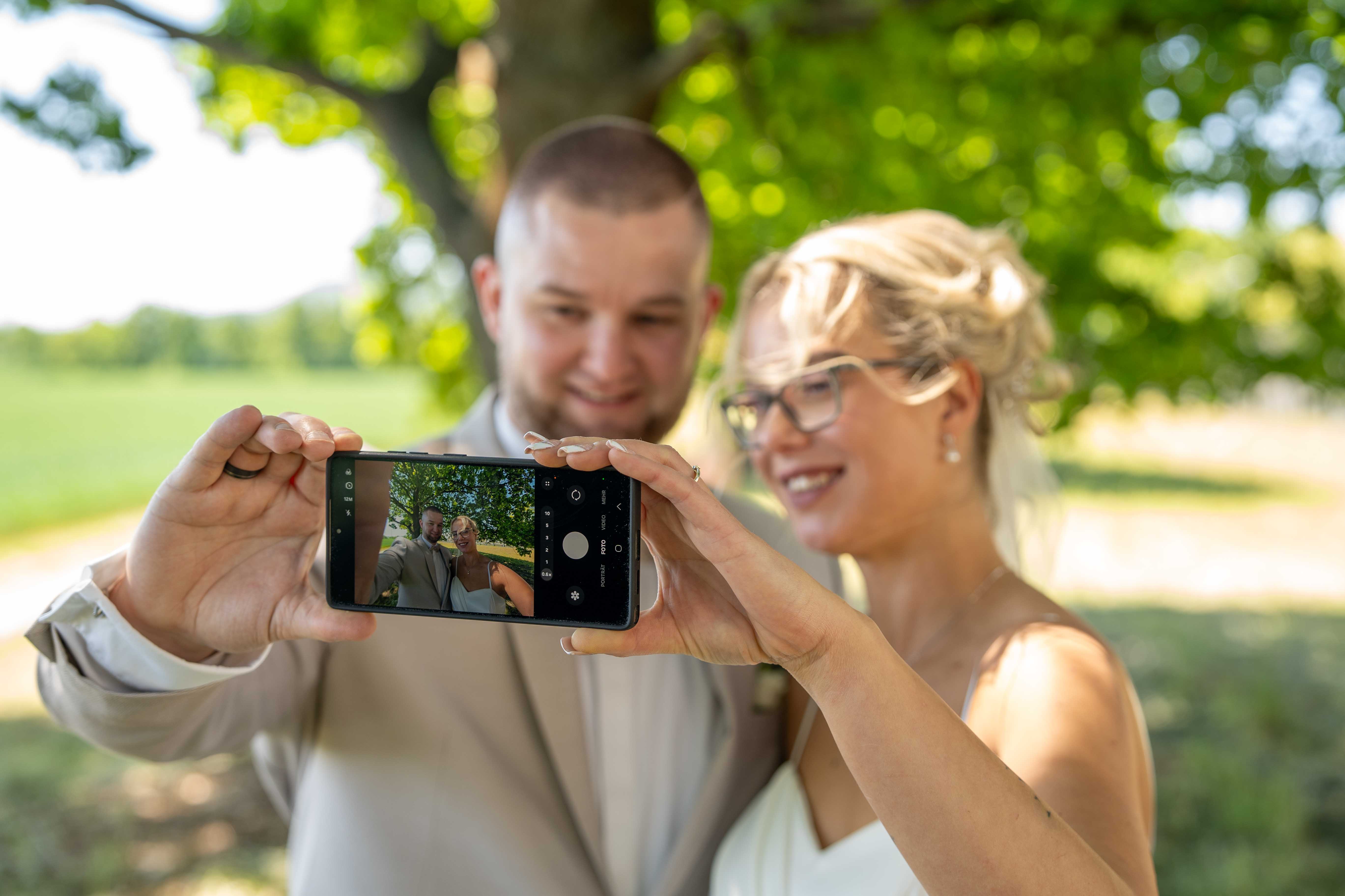Braut und Bräutigam machen bei strahlendem Sonnenschein im Freien ein fröhliches Selfie mit dem Smartphone; der Bräutigam hält das Handy.