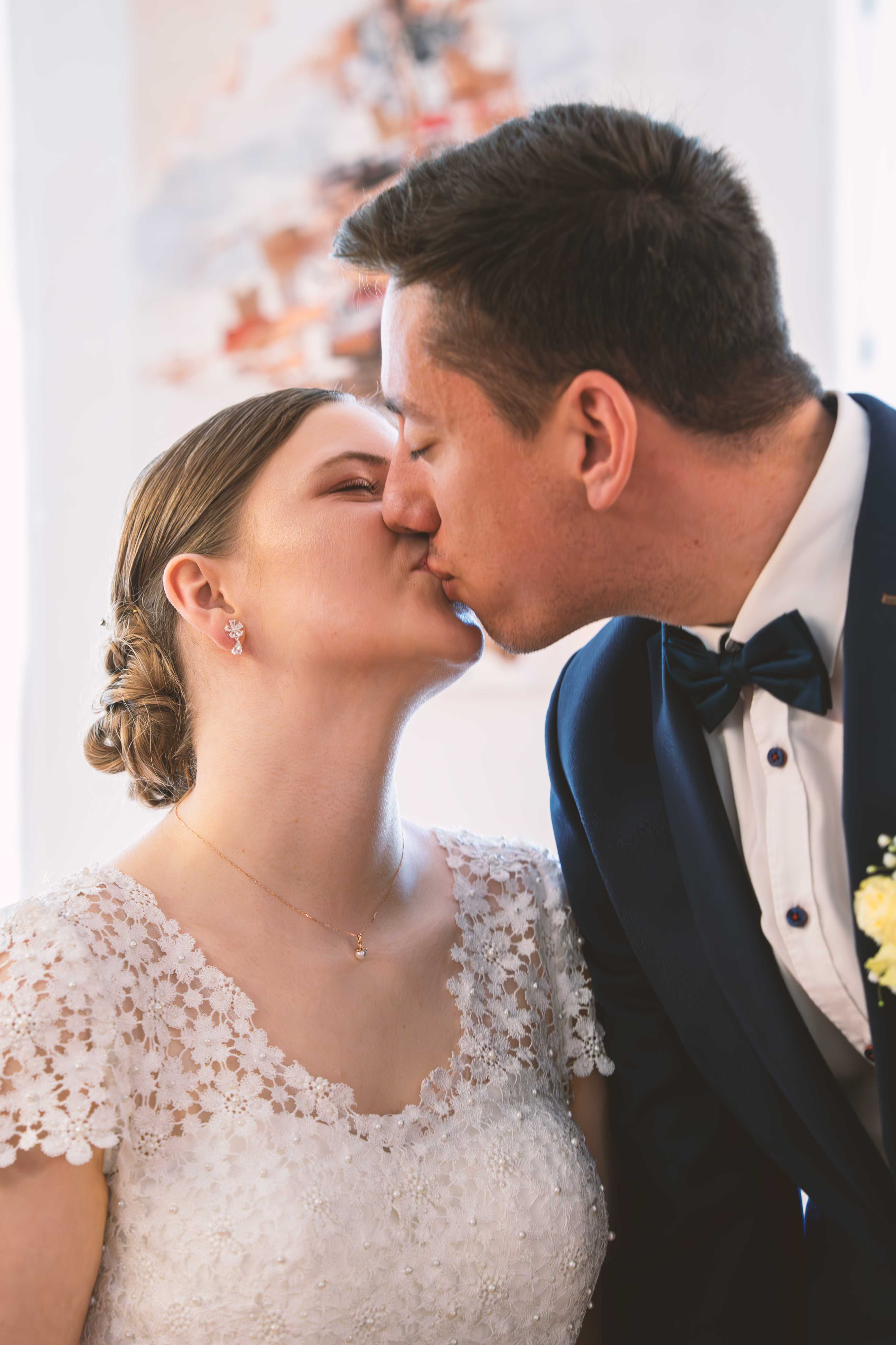 Nahaufnahme von Braut und Bräutigam beim romantischen Kuss während der standesamtlichen Trauung. Die Braut trägt ein weißes Spitzenkleid mit Perlen, der Bräutigam einen dunkelblauen Anzug mit Fliege.