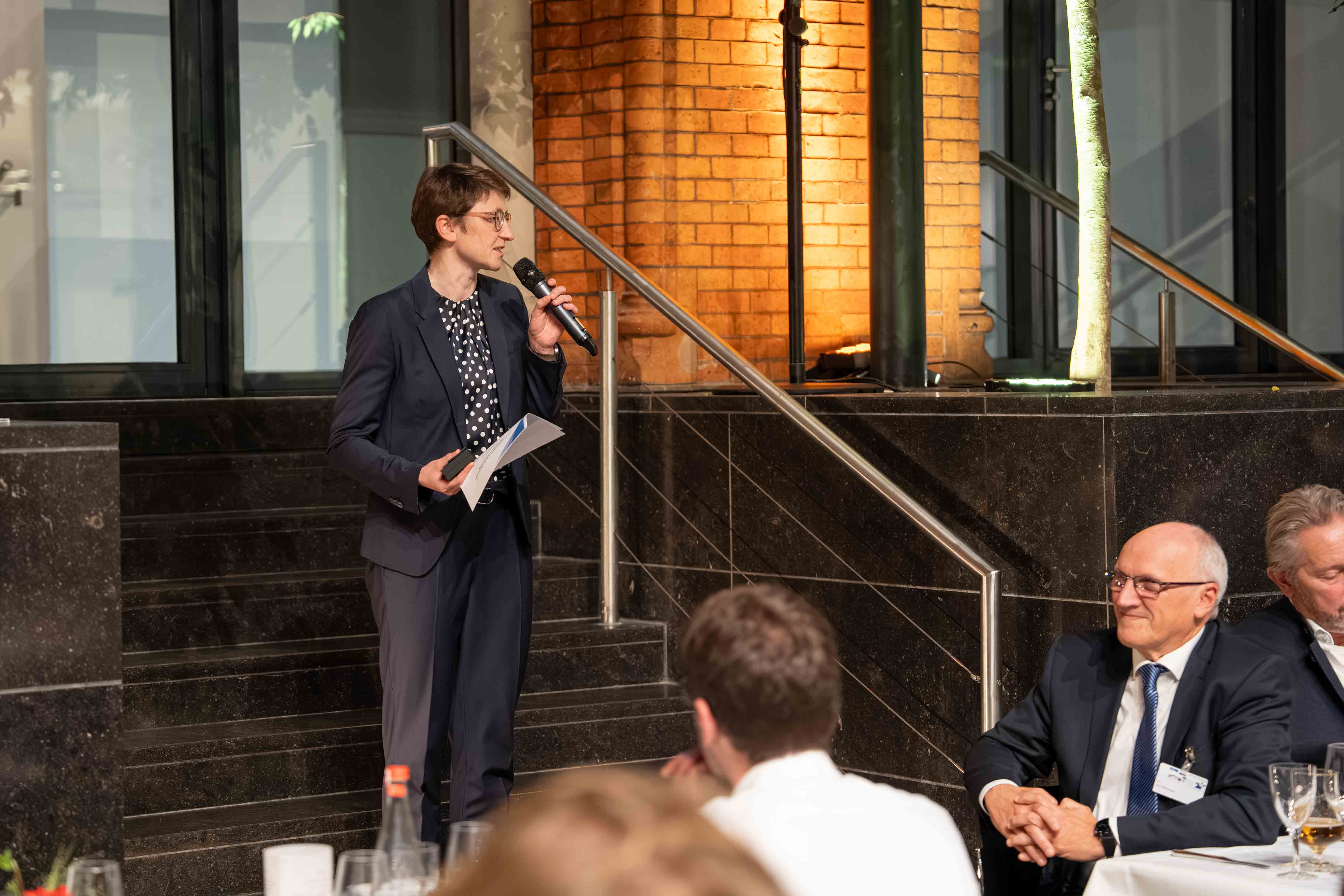 Eine Frau im Business-Anzug steht auf einer Treppe und spricht in ein Mikrofon vor sitzendem Publikum bei einem Dinner.