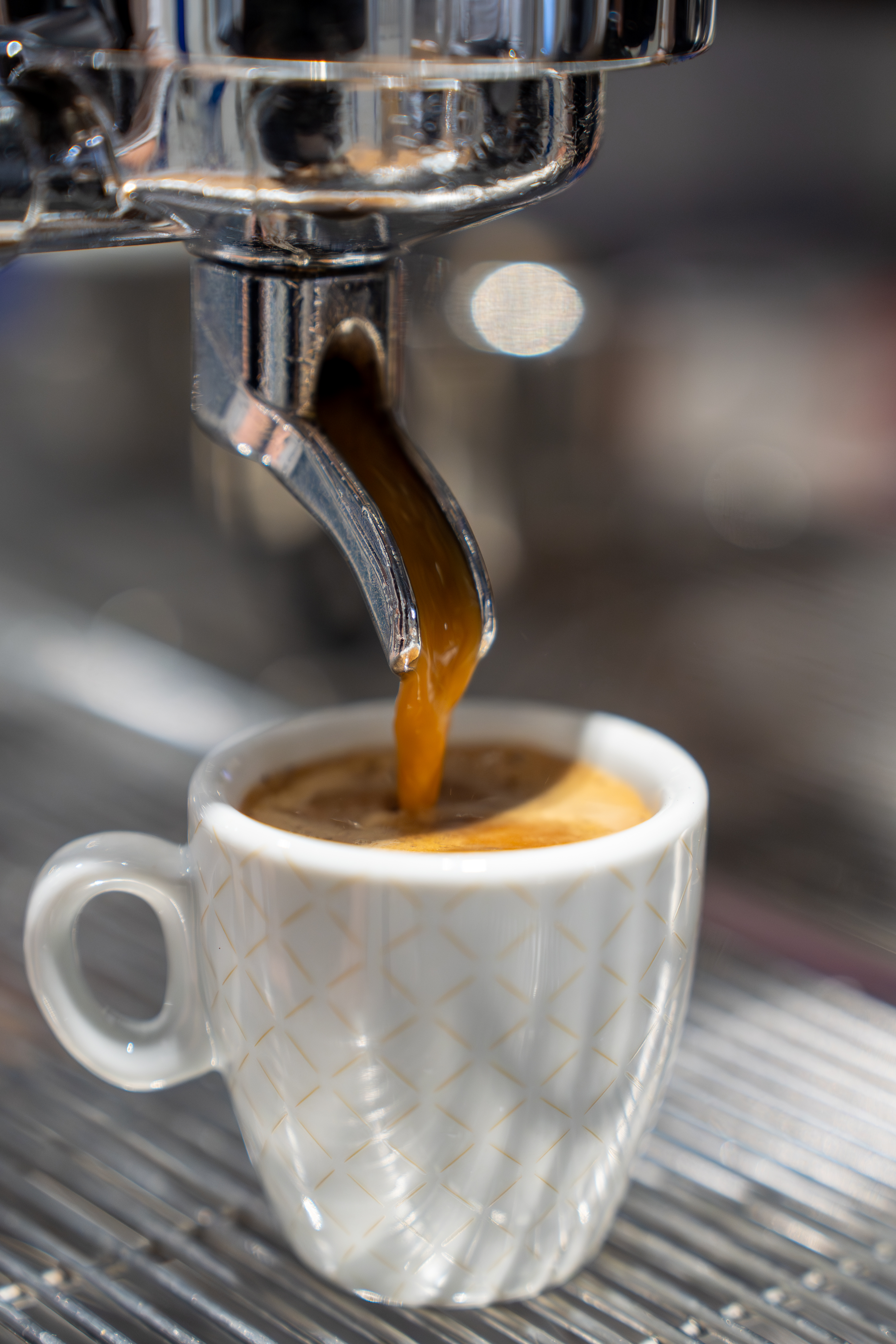 Frischer Espresso läuft aus dem Siebträger einer Kaffeemaschine in eine kleine weiße Tasse mit dezentem goldenem Rautenmuster.