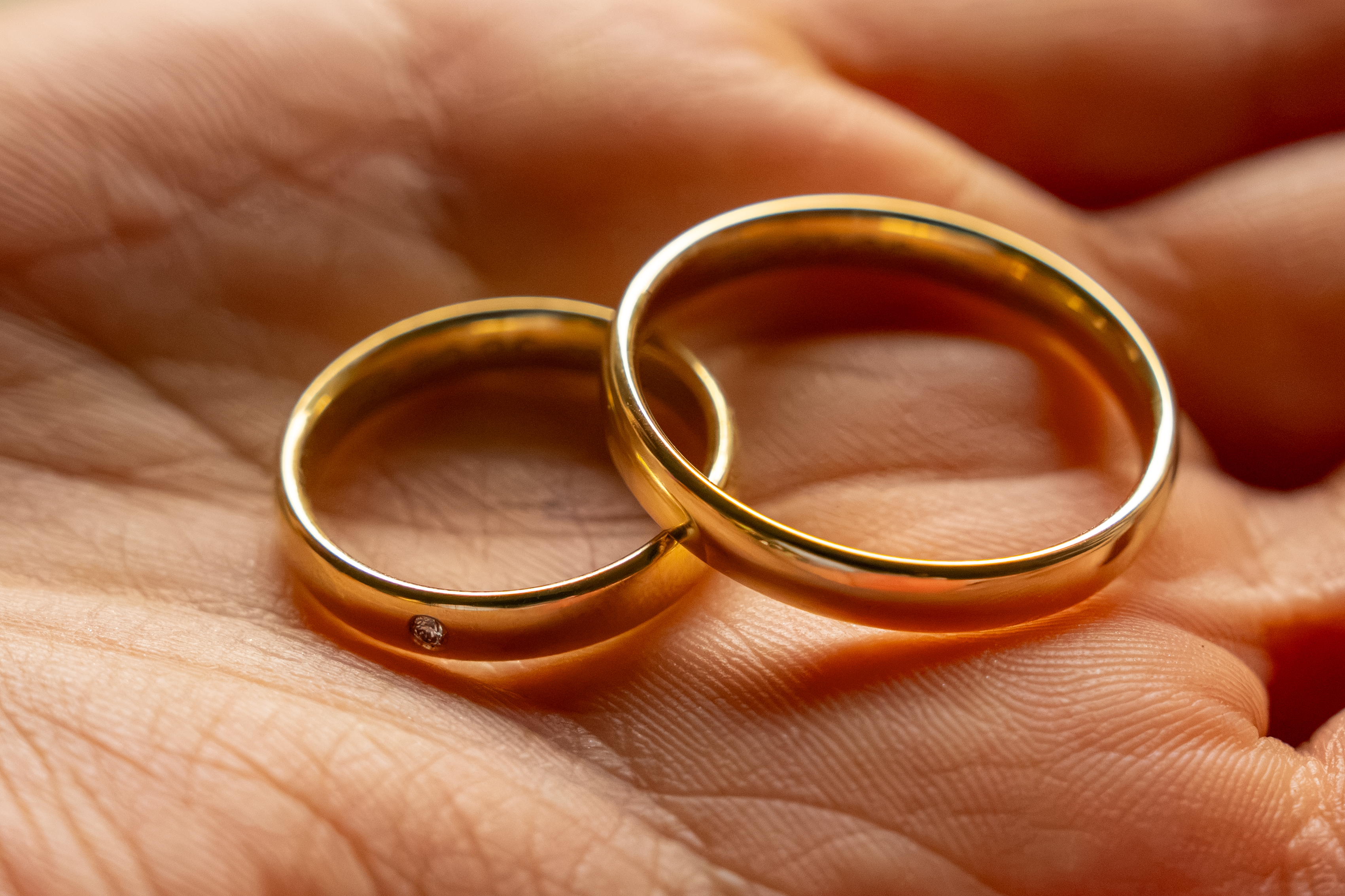 Makroaufnahme zweier ineinander verschlungener Eheringe aus Gelbgold in einer geöffneten Handfläche, einer davon mit einem kleinen Diamanten veredelt.